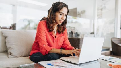person with dark mid-length hair wearing coral button up blouse sitting on a white sofa, typing on laptop on coffee table, depicting Supporting Family Caregiving report for Milken Institute