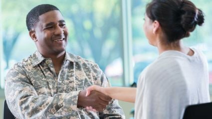 An image of a smiling man with brown skin and short brown hair, wearing camouflage military attire, shaking hands with a woman with white skin and brown hair in a bun, wearing a white shirt.