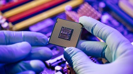 An image of hands with light purple gloves holding what appears to be a computer chip with a purple-ish background of technical computer pieces.