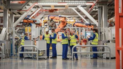 Workers in high-visibility vests and hard hats operate and monitor machinery in a modern factory with large robotic arms.