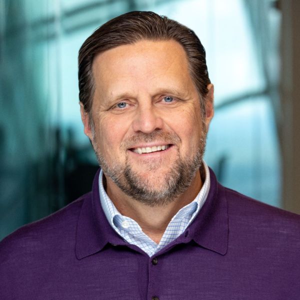 An image of a man with white skin, blue eyes, combed brown hair, grey and brown facial hair, smiling and wearing a purple shirt with a blue collar poking through.