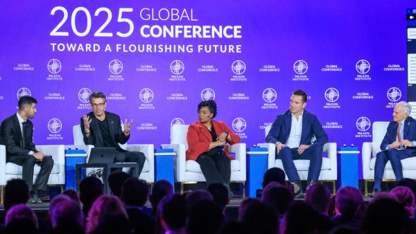 Panel of five speakers seated on stage at the 2025 Milken Institute Global Conference, with a vibrant purple backdrop displaying the event theme 'Toward a Flourishing Future,' as audience members watch.