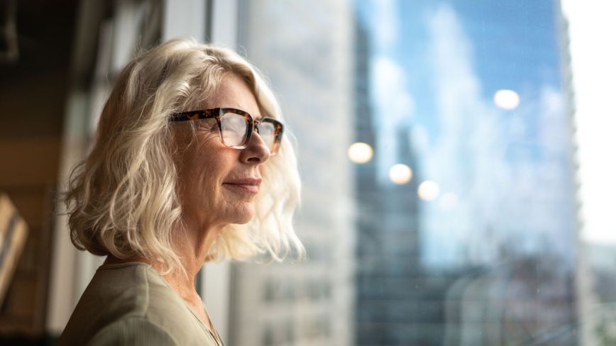An older woman with blonde hair looks out a window of a highrise