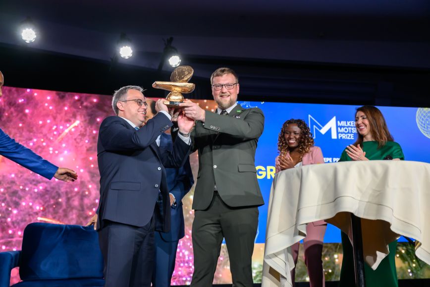 Two light-skinned males receive an award on stage at an event as two women, one Black and one white applaud for them