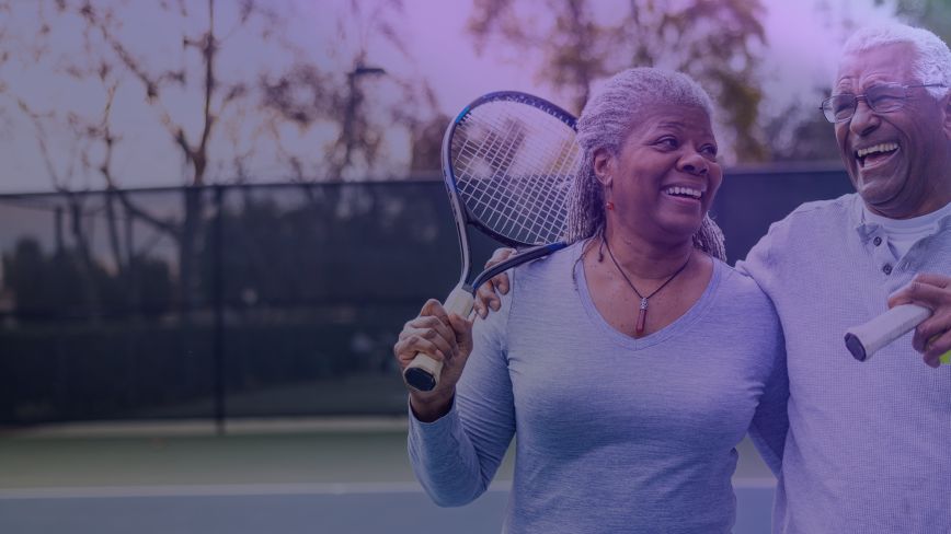 An older man and woman laugh with arms around each other while playing tennis