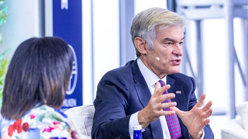 A man identified as Mehmet Oz, wearing a navy suit, white shirt, and patterned pink tie, speaks animatedly into a headset microphone, gesturing with both hands. He is seated on stage in conversation with a woman in a colorful floral blouse, whose back is to the camera. The background shows a blurred event setting with signage.