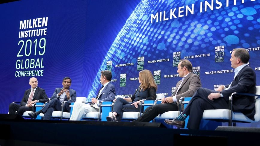 A panel of six speakers sits on stage at the Milken Institute 2019 Global Conference, with a large digital backdrop displaying the conference name and logo.