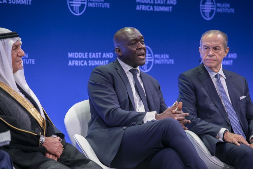 Three men sit on stage at the Milken Institute Middle East and Africa Summit. The man on the left wears traditional Middle Eastern attire, the man in the center wears a dark suit and speaks while gesturing with his hands, and the man on the right, also in a dark suit, listens attentively.