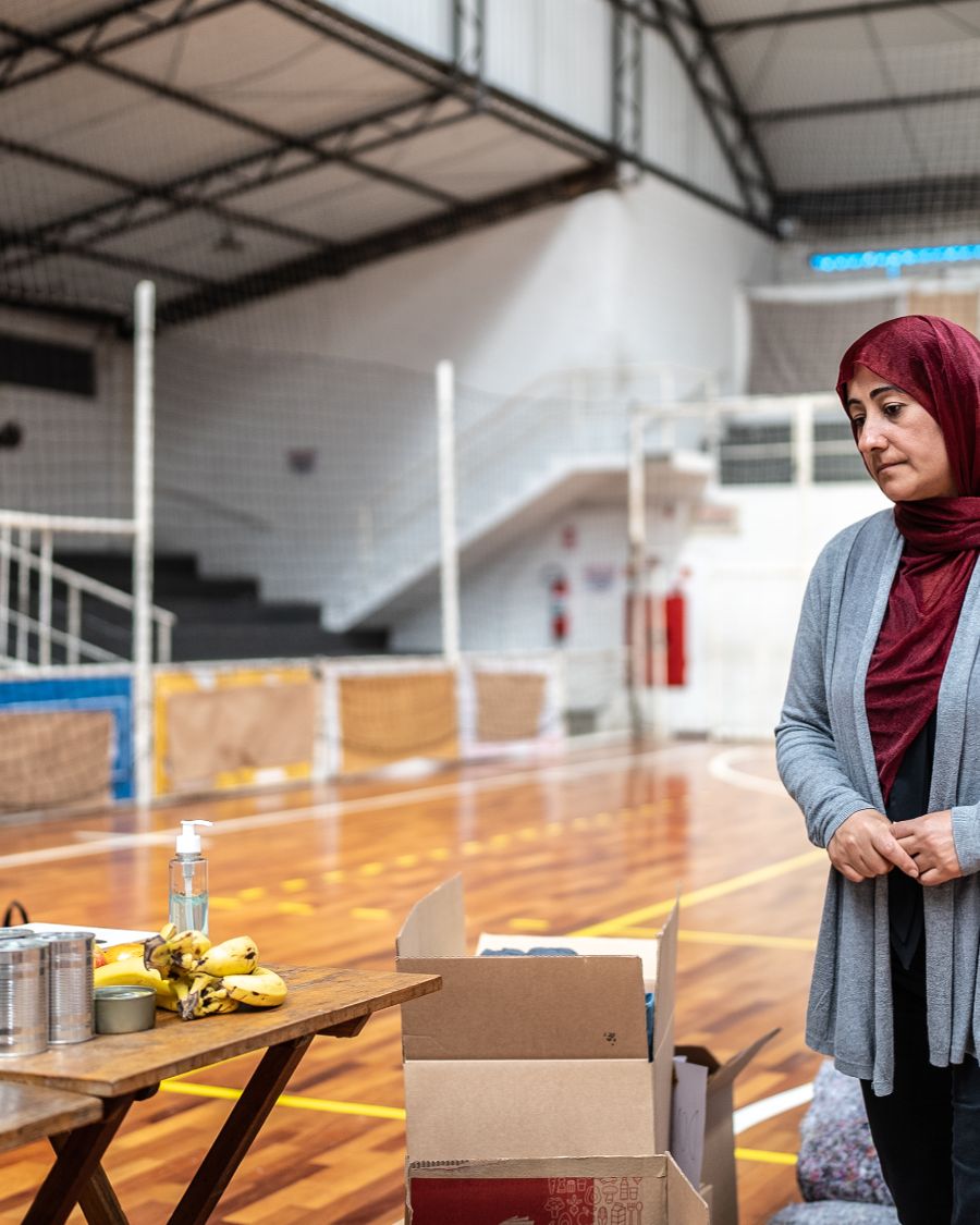 An image of various adults standing in a line in what appears to be a school gymnasium and waiting to collect food from a table, indicating a food bank or food drive.