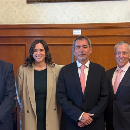 An image of the Milken Institute's CEO, Richard Ditizio, standing in front of a flag of Mexico beside four people. Rich has white skin and gray hair and is wearing a suit. There are two other men with white skin wearing suits, one has gray hair and one has black hair. There is also a woman with a tan jacket, white skin, and short brown hair.