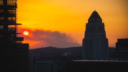 Los Angeles City Hall silhouetted at sunset as wildfire smoke drifts across the skyline, with an orange sky and the sun partially obscured.