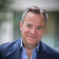 A light-skinned male-presenting person with short, dark hair wearing a suit jacket against a blurred background