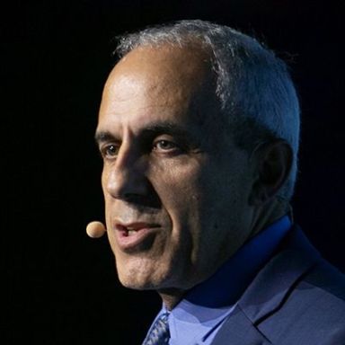 An image of an older adult man with white and slightly tan skin and short white hair. It is an image of mostly his head and he is speaking into an earpiece microphone and wearing a blue suit and tie. He is against a black background.