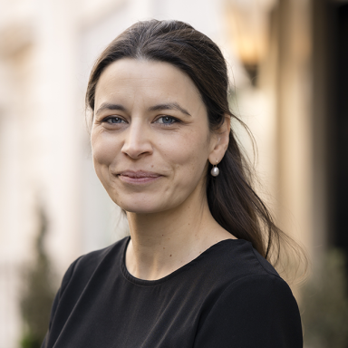 Sophie Eggar, Director of Strategic Partnerships, Europe at the Milken Institute, smiling outdoors in professional attire. She has long dark hair pulled back, wears pearl earrings, and is dressed in a black top. The background is softly blurred, suggesting a city or institutional setting.