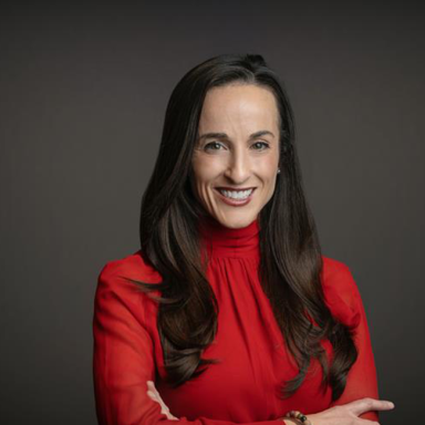 Headshot of a smiling woman with long dark brown hair, wearing a bright red blouse, standing against a dark gray background with her arms crossed.