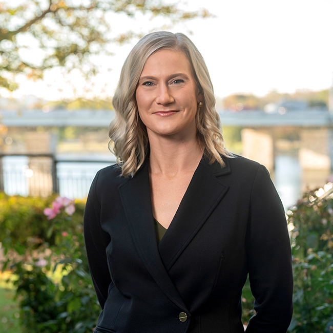 An image of a smiling light-skinned female-presenting person named Elizabeth Burke with light hair wearing a black shirt against a nature-ish background