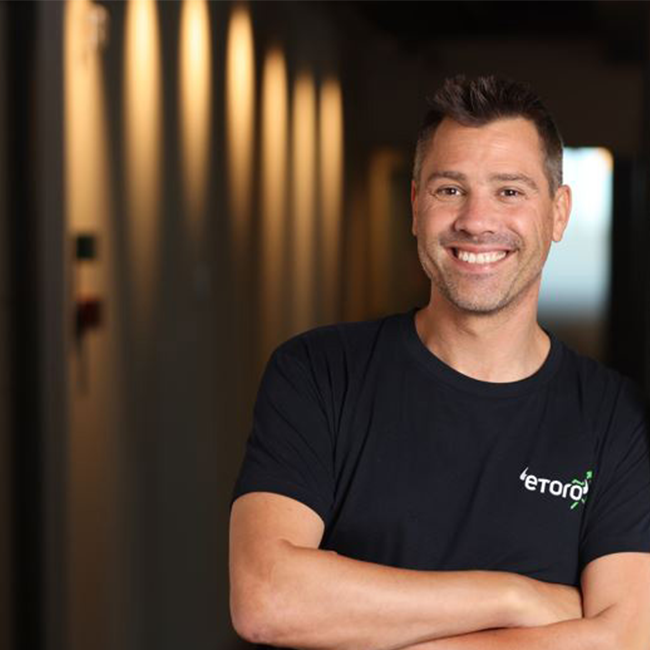 An image of a man named Yoni Assia, standing in a hallway with his arms crossed, smiling at the camera, and wearing a black t-shirt. He has white skin and short brown hair.