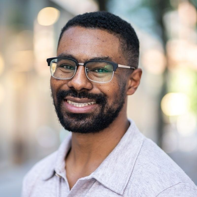 A headshot of a man named Alexander Meeks. He is standing against a blurred background and smiling at the camera. He has brown skin, a short dark brunette beard, short dark brunette hair, and brown eyes. He is wearing glasses and a gray collared polo shirt.