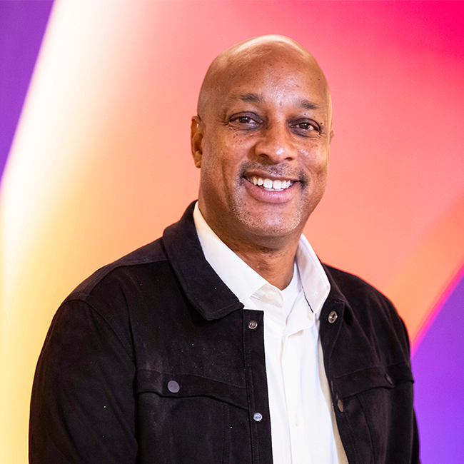 An image of a man named David Levy. He is standing against a pink and purple colorful background and smiling at the camera. He has light brown skin, a bald head, and is wearing a denim jacket over a white shirt.