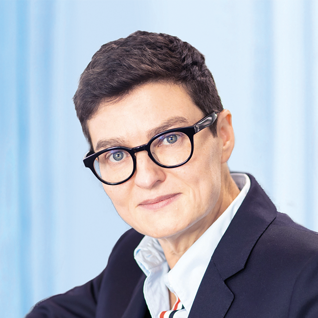 An image of a person named Robyn Grew. This person has white skin, blue eyes, short brown hair, and is looking at the camera seriously, wearing round black glasses and a suit blazer.