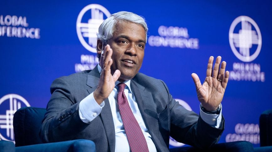 An image of an older adult man with gray hair, light brown skin, and brown eyes. His hands are raised as he is speaking on a stage and he is wearing a suit and tie. The background reads "Milken Institute" and "Global Conference." The Milken Institute's Global Conference is an annual forum that brings together leaders from business, finance, and other fields to discuss economic and social issues.